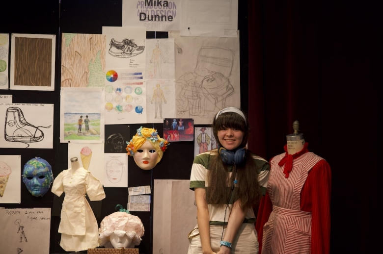 Drama: Production & Design student smiles posing with her final project, which includes sketches pinned to the wall, costumes, and a face mask.